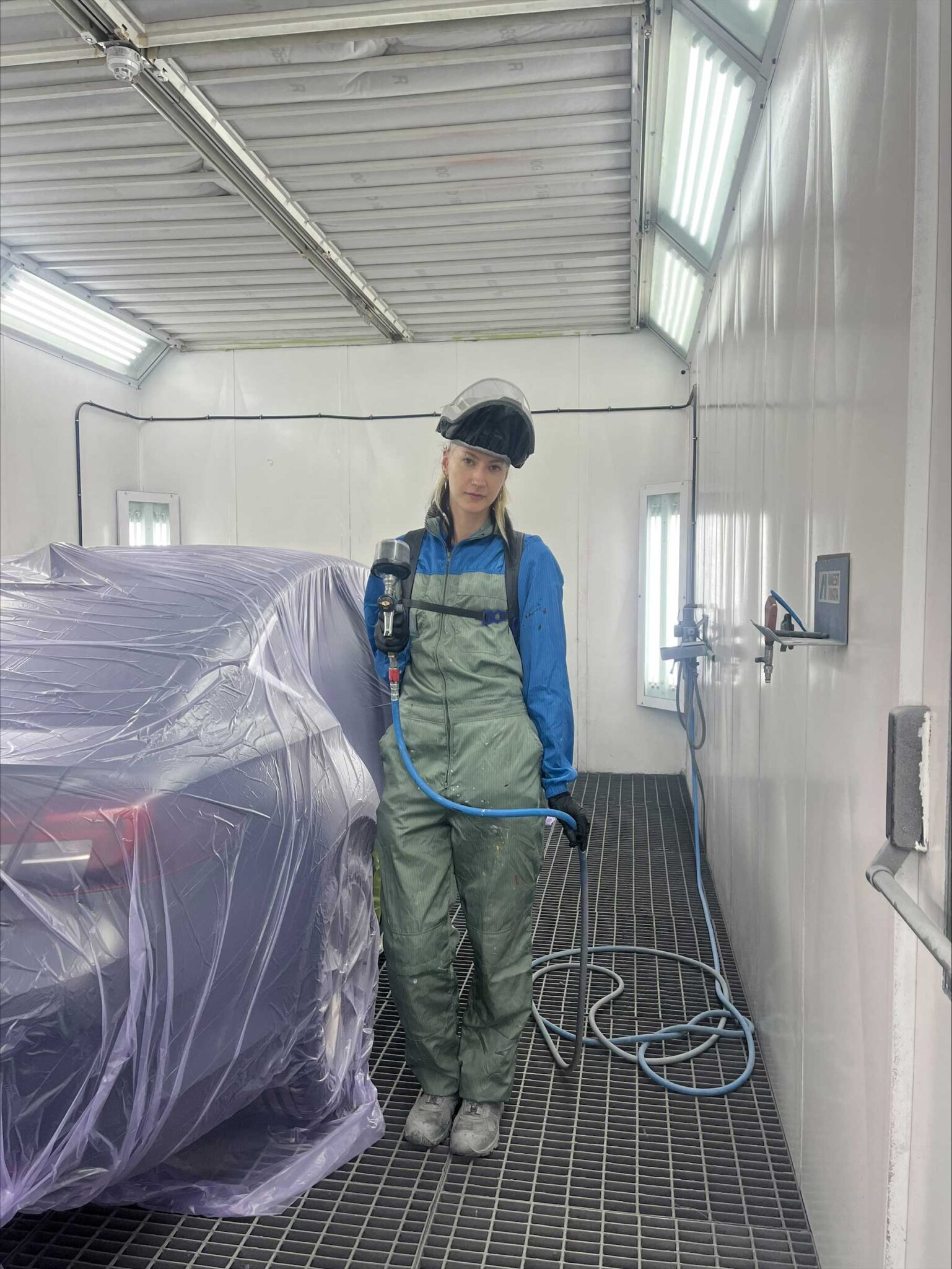 Worker in a green coverall and white helmet with face shield holding a spray gun inside a paint booth covered with plastic sheets over a vehicle.